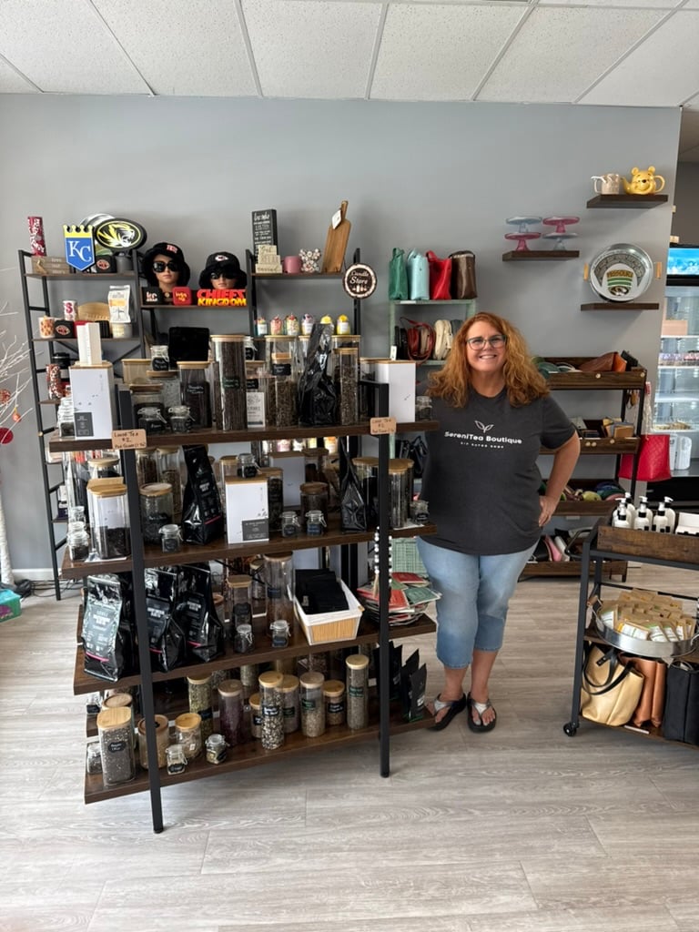 Smiling owner in a SereniTea Boutique shirt standing beside shelves of loose leaf tea jars inside the Lee's Summit shop