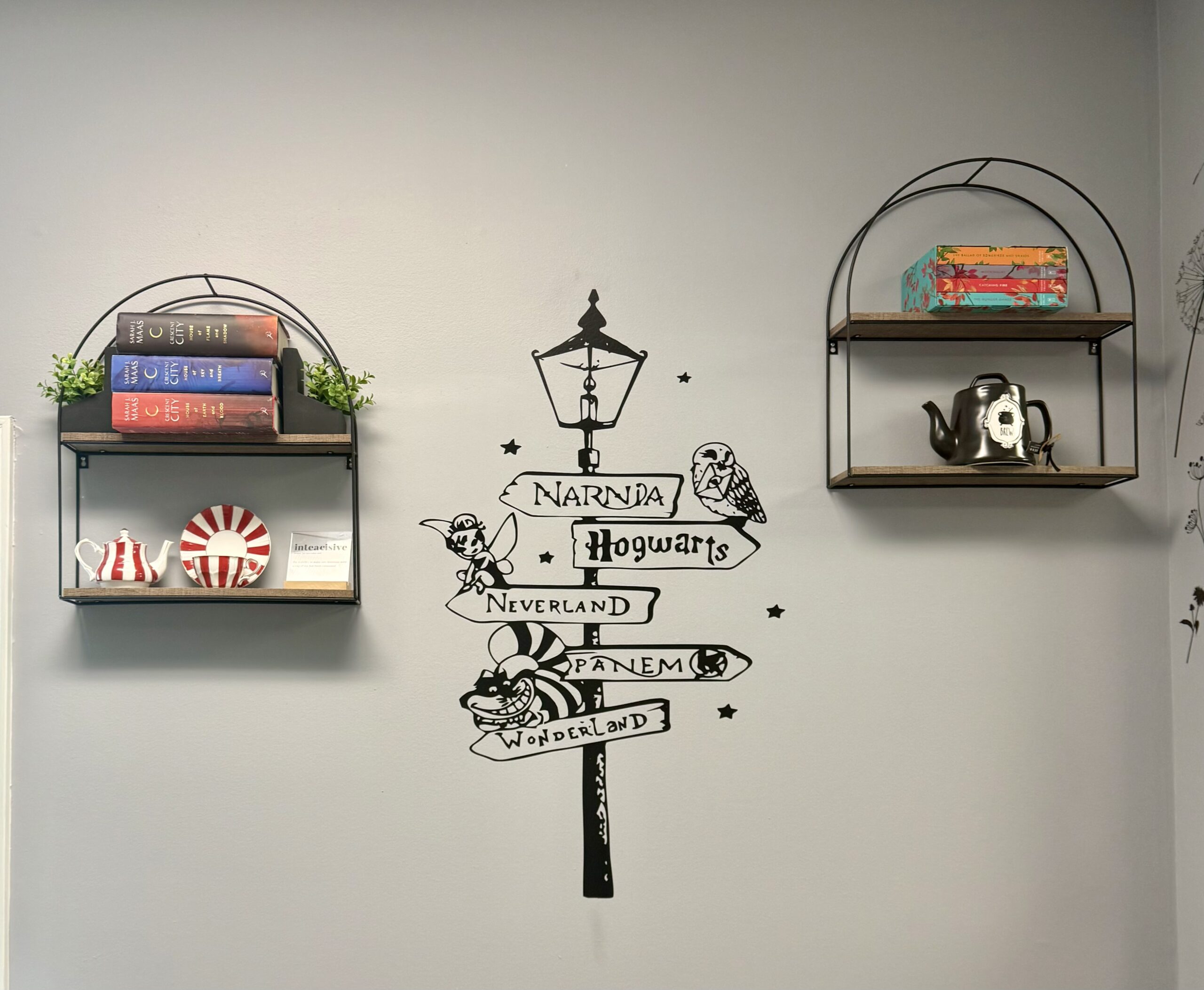 Book Nook wall at SereniTea Boutique with a signpost decal pointing to Narnia, Hogwarts, Neverland, Panem, and Wonderland between shelves of books and teapots